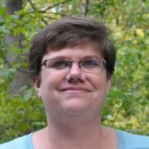 A woman with short brown hair and glasses standing in front of a wooded area